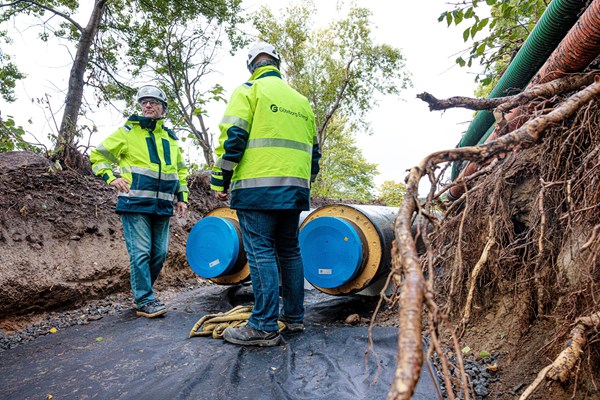 Två medarbetare i varselkläder står i vid en nedgrävd fjärrvärmeledning