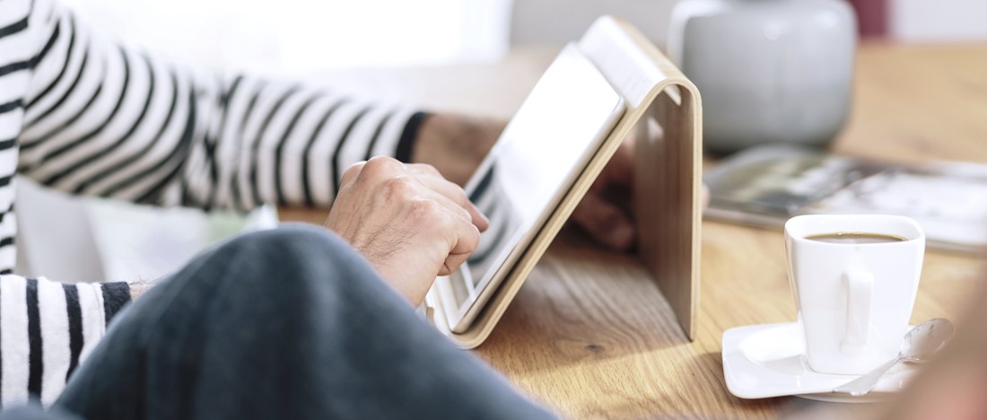 Inzoomat på en person som sitter vid ett bord i randig tröja som har en kopp kaffe och arbetar på en läsplatta