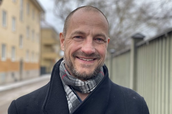 En man iklädd rock och med halsduk runt halsen ler mot kameran.