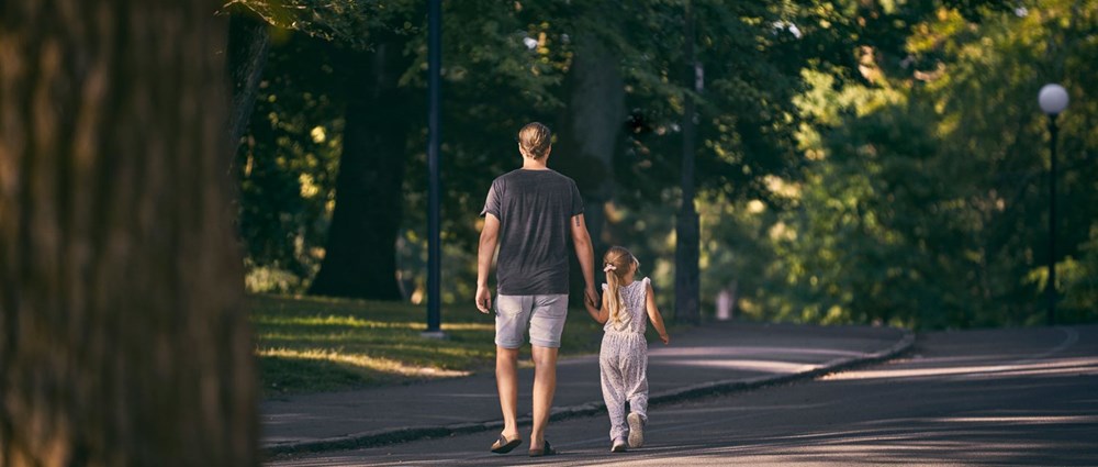 Far och dotter promenerar hand i hand genom ett somrigt Slottsskogen.
